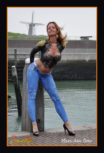 woman on a dock in a painted on detailed black vest with gold highlights and painted on jeans in front of a dutch windmill 
